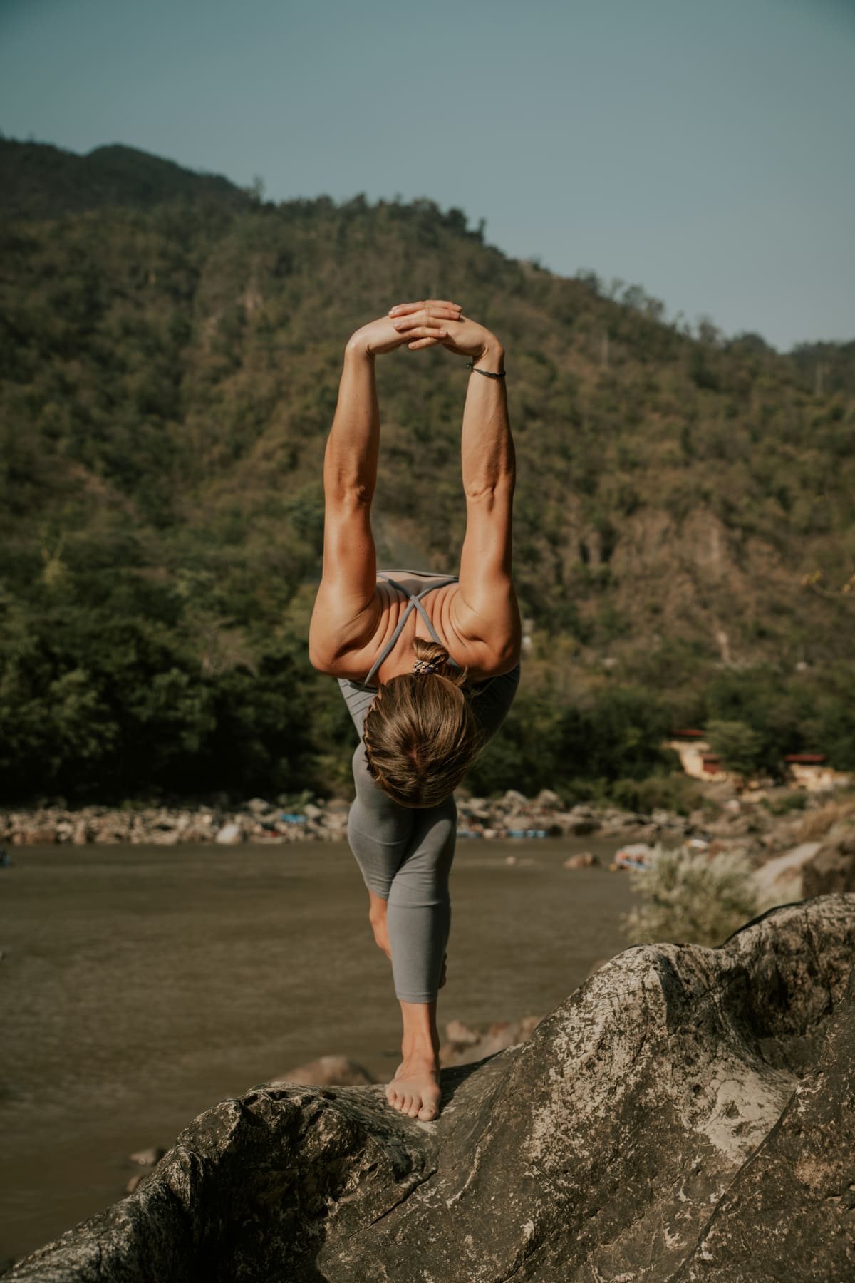 Prof de yoga en équilibre debout au bord d'une rivière de montagne, jambe levée vers le ciel